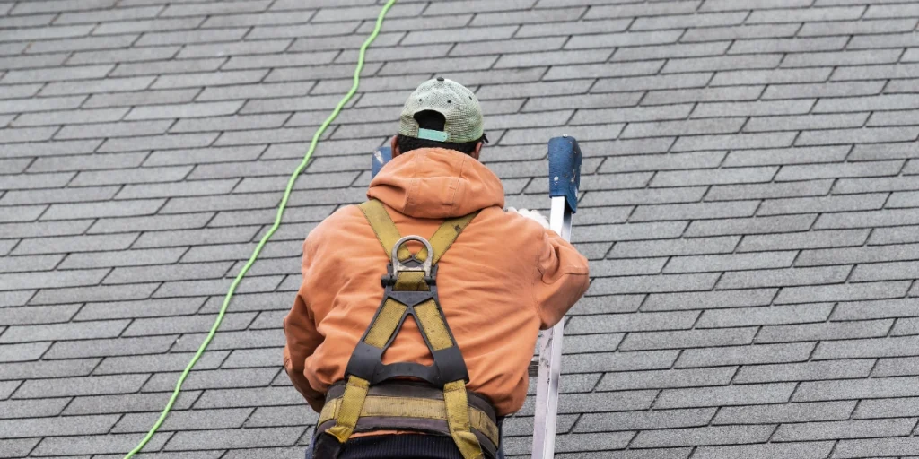 A roofing contractor in a safety harness climbs a ladder on an asphalt shingle roof during a professional inspection.