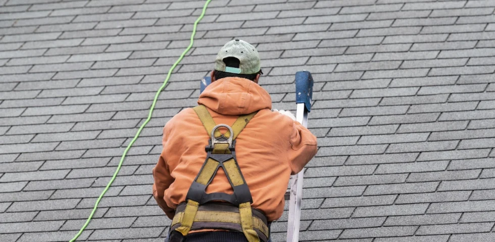 A roofing contractor in a safety harness climbs a ladder on an asphalt shingle roof during a professional inspection.