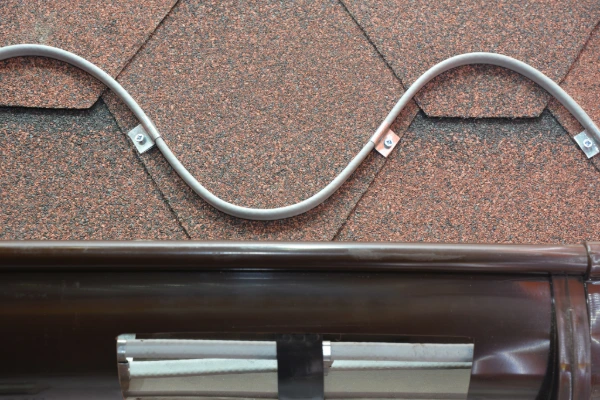 Heat cable in a zig-zag pattern secured to brown asphalt shingles above a gutter to prevent ice dam formation.