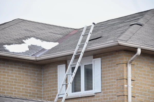 A residential home with significant shingle damage exposing the roof deck, with a ladder leaning against the exterior wall.
